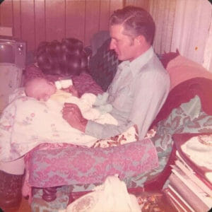 man bottle-feeding an infant in his lap in 1970's decor